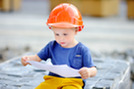 A toddler wearing an orange hard hat, looking down at a paper in deep concentration.