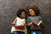 Two young girls of color lay flat on carpet looking at each other while holding books.