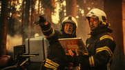 Two men in firefighter gear survey a wildfire ahead of them.