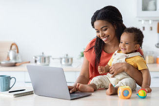 A brown mother and baby smile as they look at an open laptop in front of both of them.
