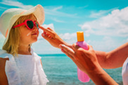 Young girl on beach wearing heart-shaped sunglasses with woman applying sunscreen on her face.