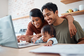 A smiling mom, dad and their curious toddler all huddle around an open laptop, on a counter top, in a brightly lit room.