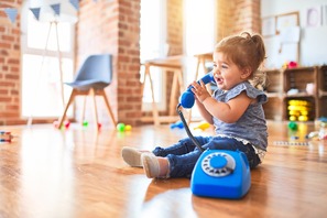 A toddler girl sitting on the floor of a sunlit playroom, surrounded by toys and holding a plastic telephone receiver to her ear with a curly cord.
