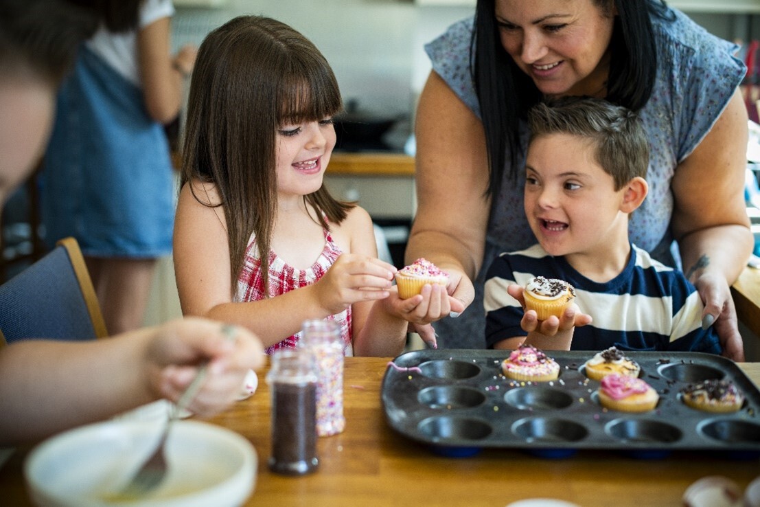 Children eat cupcakes with woman with dark hair helping them.