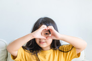 Young girl of color with dark hair has a smile on her face while she holds up both hands to her eye and makes circle around her eye.