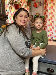 Woman with long brown hair and wearing a gray top, holds close a young girl with brown hair, green shirt, sitting on kitchen countertop.