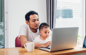 Photo of a baby sitting on their father's lap, both looking intently at a laptop.