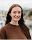 Photo of Mattea Beeson smiling with long, shoulder-length, brown hair, and wearing a brown sweater, standing in front of a blurred outside background.