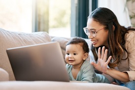 A young mother with her baby sits on the couch, as the mother waves at an open laptop screen and the baby smiles.