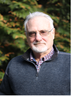 Medium shot of Greg Abell smiling, in front of a tree-filled setting in the background, wearing a grey half-zip sweater and plaid button up shirt.