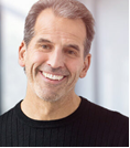 Close up photo of Doug Nathan, smiling at the camera, wearing a black sweater, in front a brightly lit background.