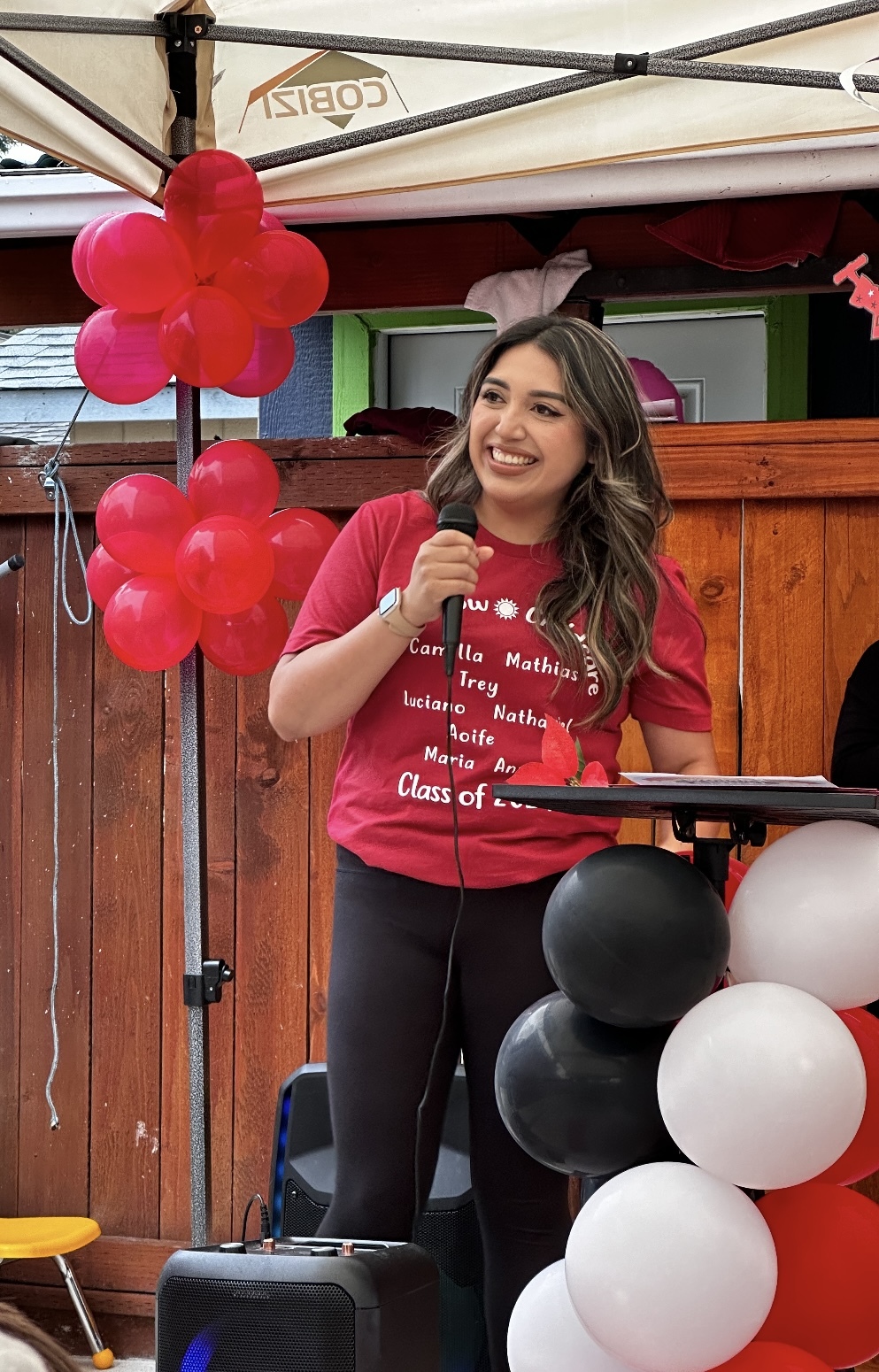 Woman with dark hair and red shirt speaks into microphone with wooden fence behind her.