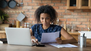 Woman of color sits at kitchen table focusing on laptop and budgeting.