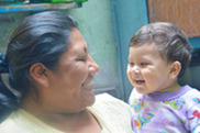 Baby girl with dark hair smiles in arms of woman of color with dark hair.