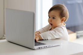 Baby sitting in front of a laptop looking surprised. 