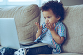 A toddler, with dark curly hair, sits on a couch looking intently at an open laptop on their lap, with both palms raised as if saying, "I don't know!"