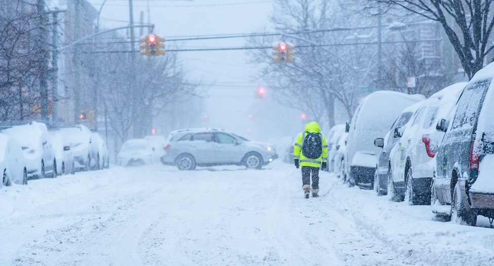Snow covered street, cars covered in snow, man walking in street, traffic stopped in distance.