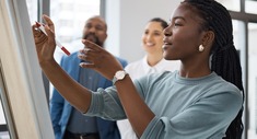 Black woman writing on white board, 2 co-workers in background watching. 