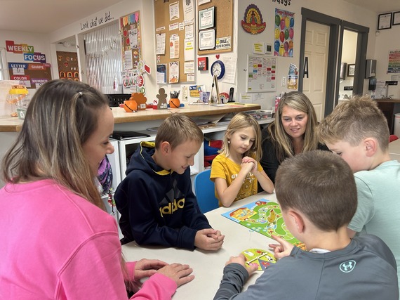 Four children sit around classroom table playing board game with two female adults.