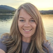 Woman with brownish blond shoulder-length hair, grayish hazel eyes, smiles with lake setting in background.