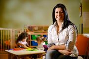 Woman of color with dark, long hair, sits in preschool classroom with legs crossed, smiling, with two seated girls in background.