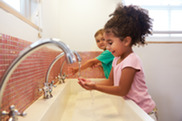 Girl with curly dark hair, pink shirt and boy with green shirt, wash hands at sink.