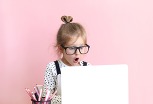 A little blonde girl with a top bun and big glasses, looks surprised, sitting behind of an open laptop, in front of a pink wall.