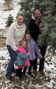 Photo of Kirsten Dixon smiling and standing next to her two young daughters, and husband, as he holds a freshly cut holiday tree in a snowy lot.