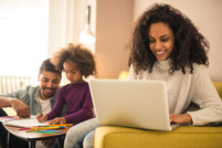 A smiling mother works on her laptop in the foreground, child and dad are visible playing in the background of the sunlit room.