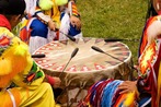 Close up shot of three Native American men in in a circle, wearing traditional regalia, playing a large a drum.