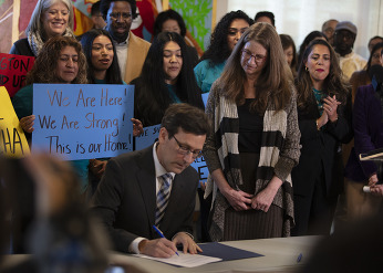 governor bob ferguson and DCYF secretary Tana Senn signing document, people with signs behind them. 