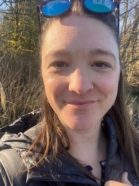 Sunlit, close shot of Alison Lofgren, smiling, wearing a warm, black jacket and sunglasses on her head, with trees and long grass in the background. 