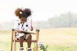 A little girl in glasses, curly pigtails and black and white sun dress, holds a red pencil, sits outside at a little desk, looking down at a notebook.