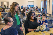 Woman stands smiling and laughing with two women sitting at table with child on top of table smiling.