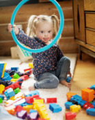 A toddler girl in blonde pigtails, sits on the floor, holding up a turquoise hoop, surrounded by colorful blocks and toys.