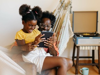 Dad and daughter relaxing together in hammock, laughing and watching a video on a tablet