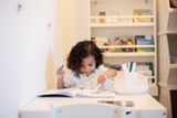 A little boy with dark, curly, shoulder-length hair and white shirt concentrates while happily coloring with markers in a coloring book.