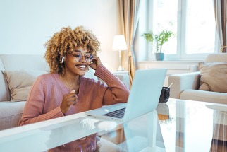 Smiling African American Woman Wearing Glasses and Wireless Earphones Makes a Video Call on her Laptop Computer at her Home Office.