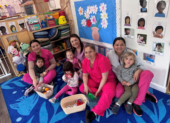 Four women and four children sit together, smiling, on carpet in classroom