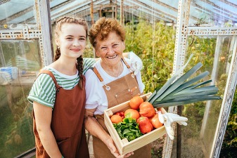grandma and teen grandaughter with box of veggies outside greenhouse