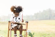 A little girl in glasses, curly pigtails and black and white sun dress, holds a red pencil, sits outside at a little desk, looking down at a notebook.