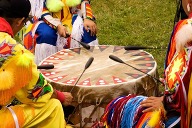 Close up shot of three Native American men in a circle, wearing traditional regalia, playing a large a drum.