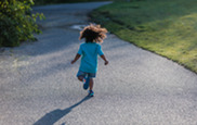 A toddler with dark curly, shoulder-length hair, a turquoise t-shirt and blue jean shorts, happily runs away from the camera and down a sunny street.