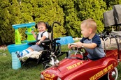 Two boys playing outside in a yard, one sits in a toy car and the other sits in a wheelchair next to an interactive play set. 