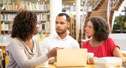 Two women and one man sit at table with laptop in library.