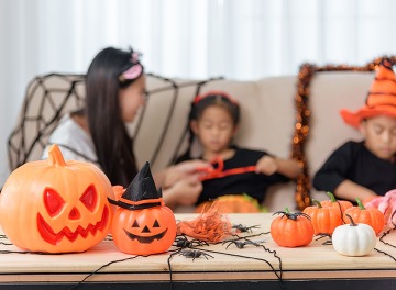 Pumpkins with smiling faces and witch hats decorate a table, with children in costumes visible in the background celebrating halloween