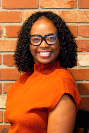 Medium shot of Tutrecia Baker, smiling, wearing black-framed glasses and an orange top, standing in front of a brick background.