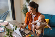Woman holding baby sitting at desk working on computer.