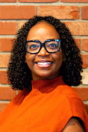Tutrecia Baker, smiling, wearing black-framed glasses and an orange top, standing in front of a brick background.