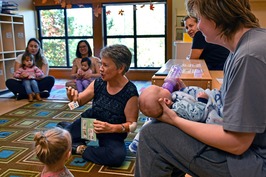 A group of mothers holding their babies, in a class setting, sit attentively around a teacher at the Hands On Children's Museum.
